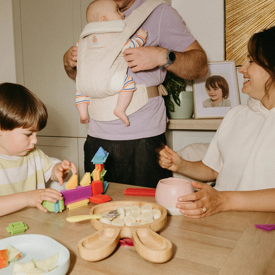 Un homme tenant un bébé dans un porte-bébé beige, regardant un enfant en bas âge qui joue à table avec une femme