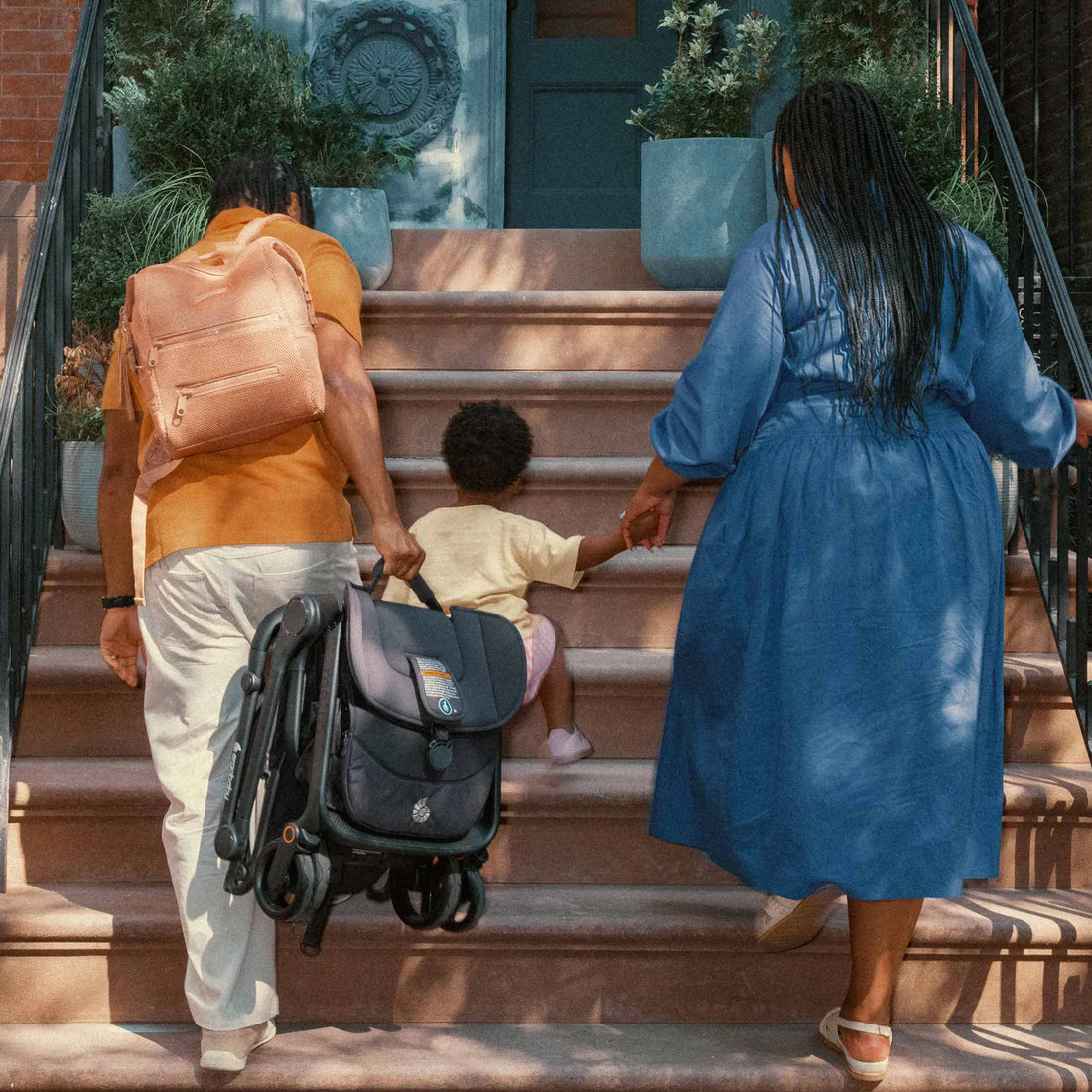 Eine Frau, die Händchen mit einem Kleinkind hält, steigt die Treppe hinauf, zusammen mit einem Mann, der einen Rucksack und einen Reisekinderwagen trägt