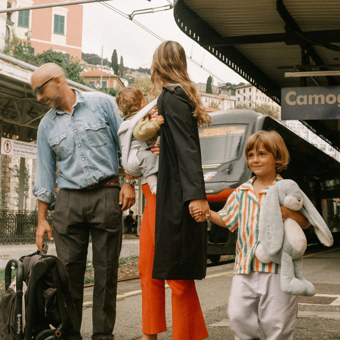 Eine Frau, die ein Baby in einer Babytrage hält und Händchen mit einem Kleinkind Babytrage , steht neben einem Mann, der einen Reisekinderwagen trägt