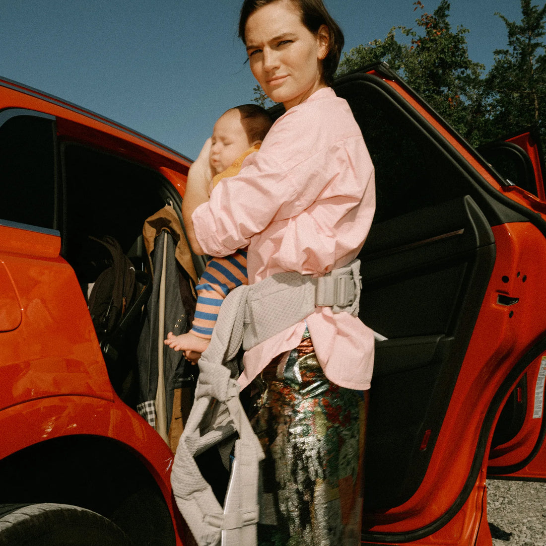 Eine Frau, die ein Baby in einer grauen Babytrage hält und vor einem Auto Babytrage