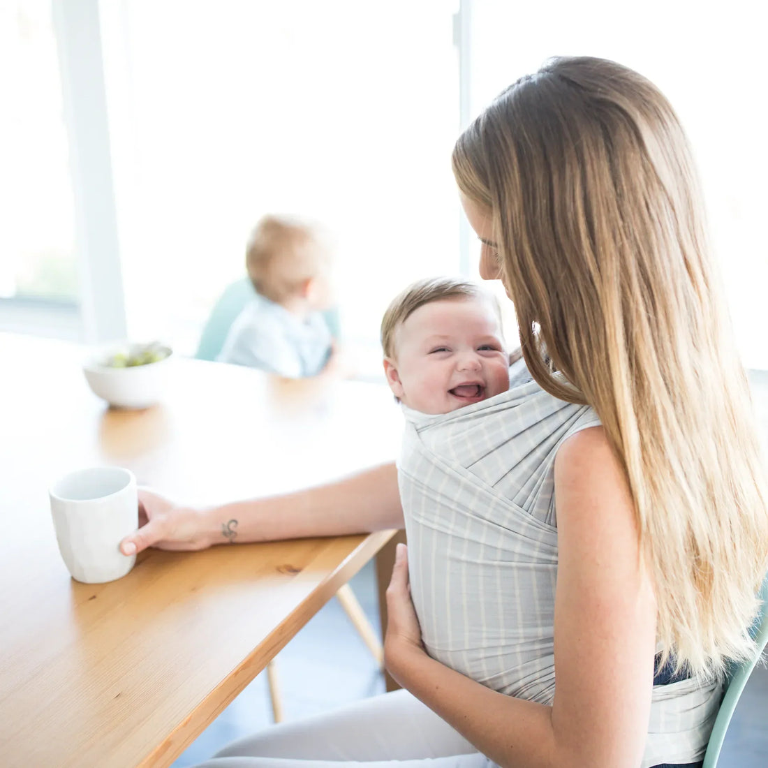 Eine Frau, die ein Baby in einem grau gestreiften Tragetuch hält, während sie an einem Tisch sitzt