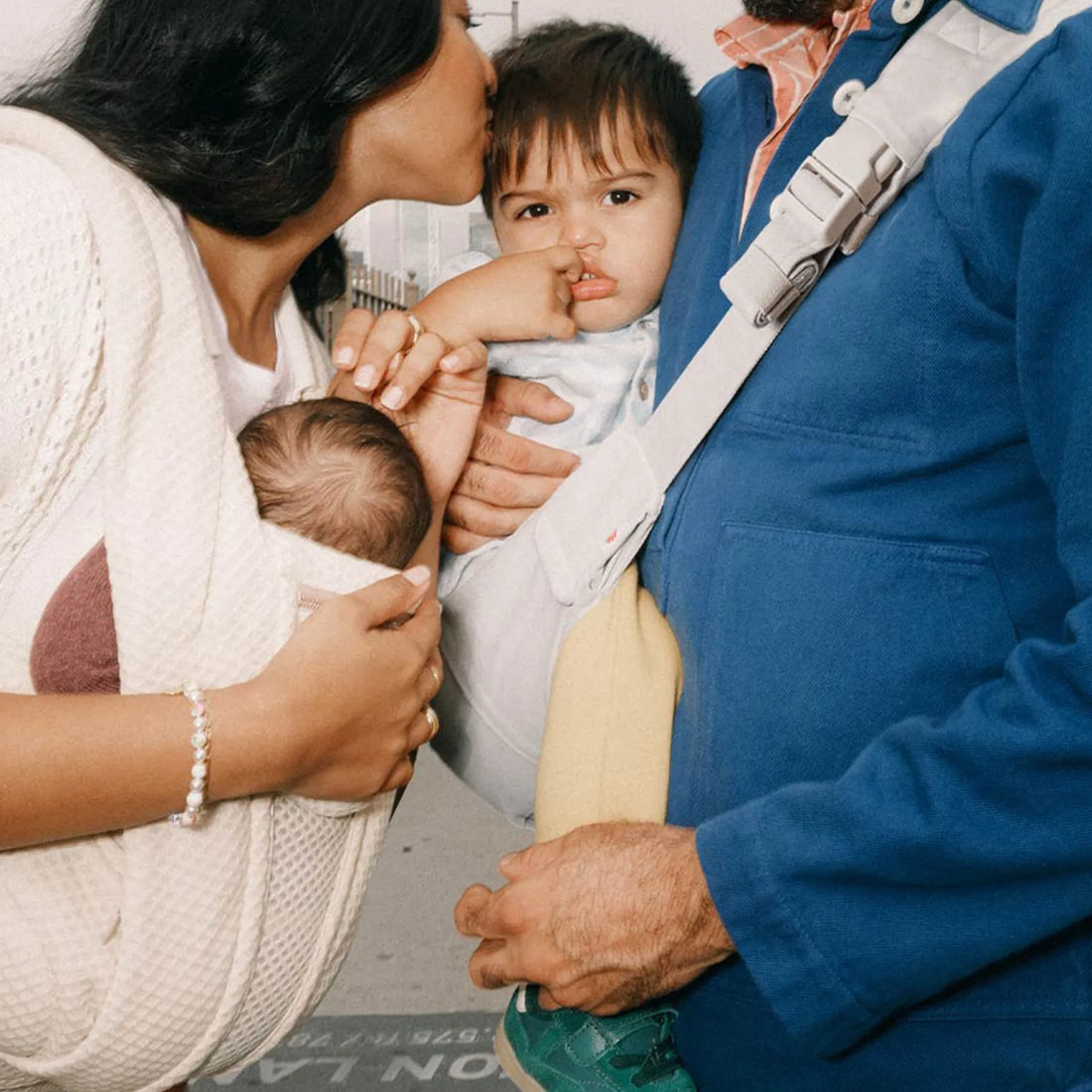 Eine Frau, die ein Baby in einer beigen Babytrage hält, Babytrage einem Mann, der ein Kleinkind in einem grauen Tragetuch hält