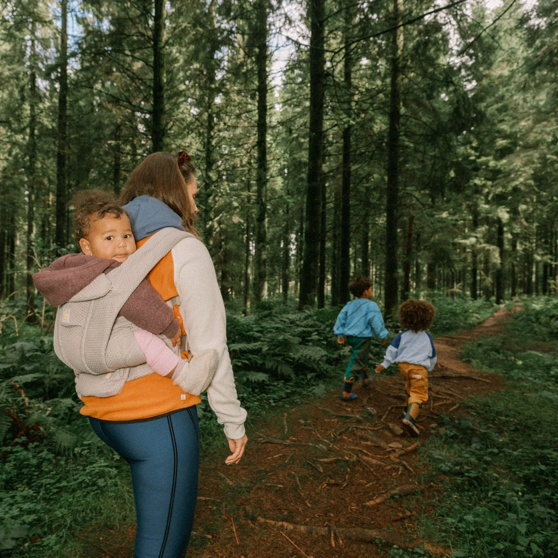 Eine Frau, die ein Baby in einer beigen Babytrage auf dem Rücken trägt und zwei Kleinkindern Babytrage