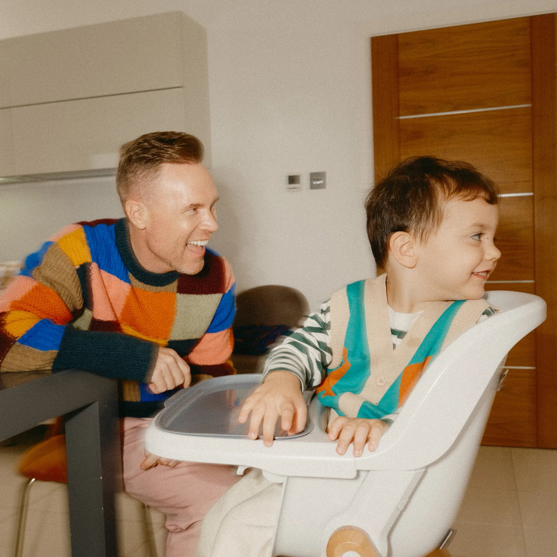Ein Mann sitzt an einem Tisch, neben ihm sitzt ein Baby im Hochstuhl, und er lacht