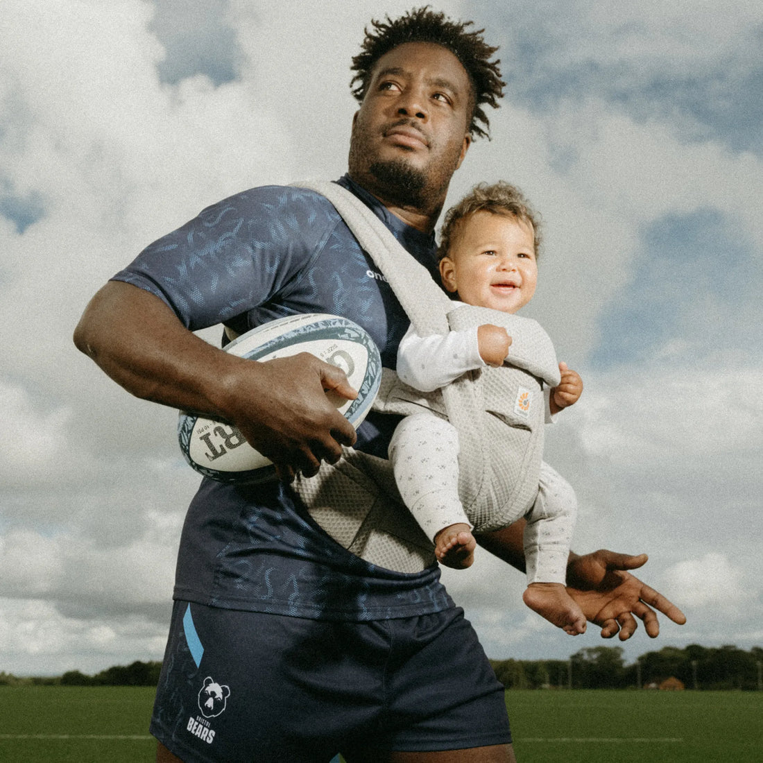 Ein Mann, der ein Baby in einem grauen Babytrage einen Fußball hält