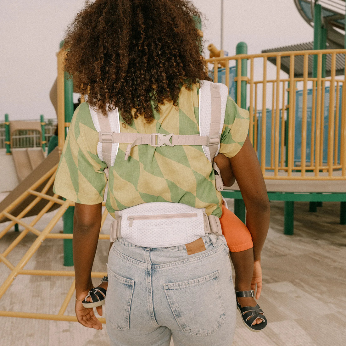 Baksidan av en person som håller ett barn i en bärsele på en lekplats