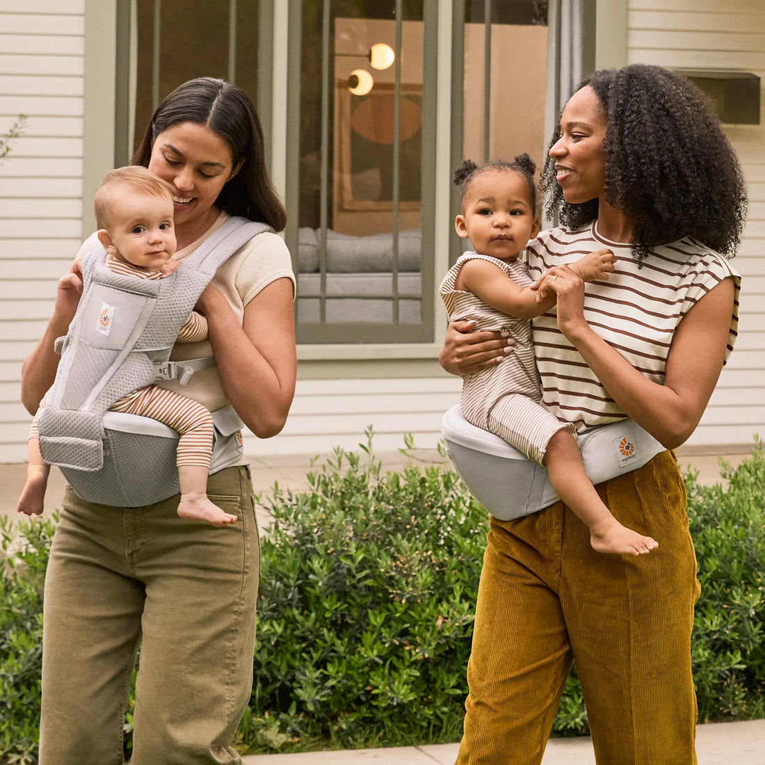 Eine Frau, die ein Baby in einer grauen Babytrage trägt, Babytrage neben einer Frau Babytrage , die ein Baby in einer Hüfttrage auf der Hüfte trägt