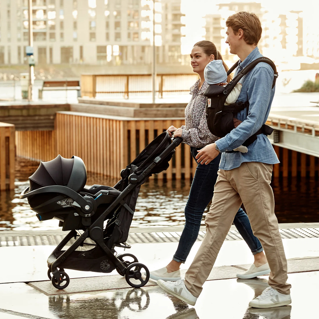 Ein Mann, der ein Baby in einer schwarzen Babytrage hält, Babytrage neben einer Frau Babytrage , die einen Kinderwagen schiebt