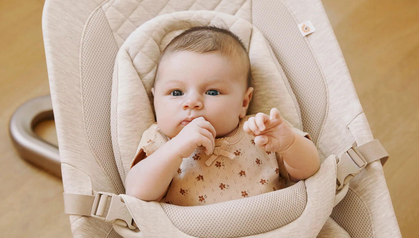 Ein Baby sitzt in einem beigen Kindersitz vor einem neutralen Hintergrund