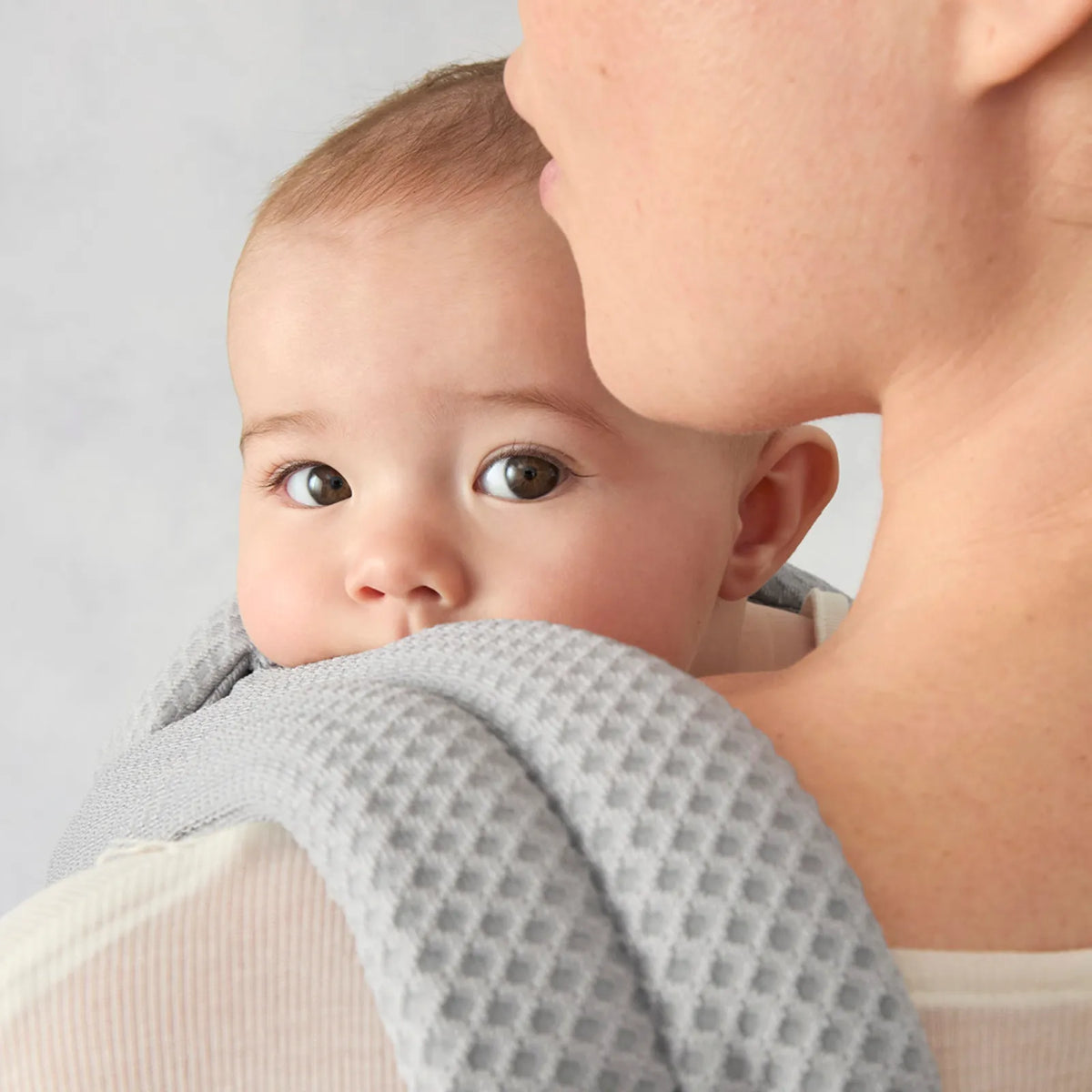 Ein Baby, das von einer Person gehalten wird, die ein graues, strukturiertes Kleidungsstück trägt, vor einem hellen Hintergrund
