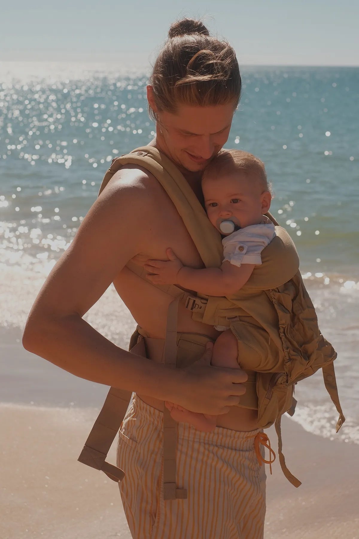 Ein Vater trägt sein Baby inBabytrage  omni classic Babytrage Strand, im Hintergrund ist das Meer zu sehen