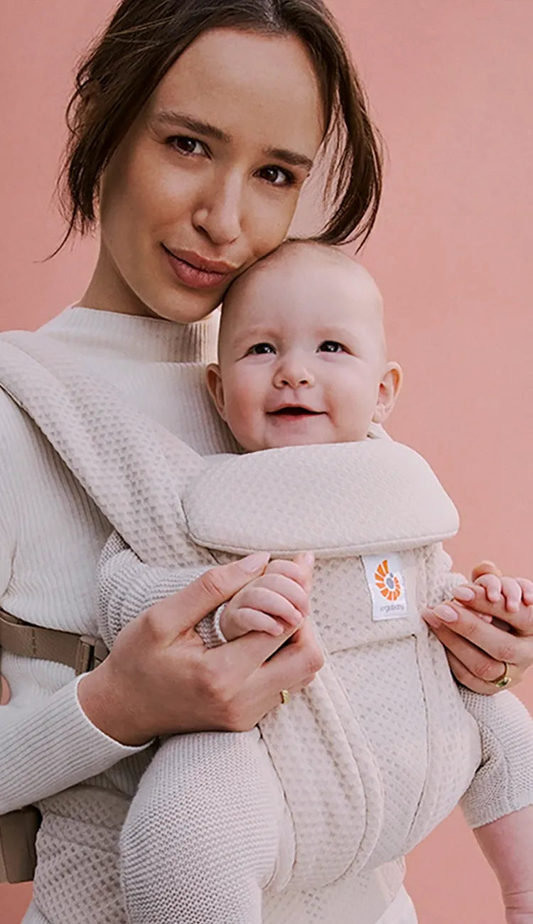 Eine Mutter, die ihr Baby in einer Ergobaby omni deluxe vor einem rosa Hintergrund hält