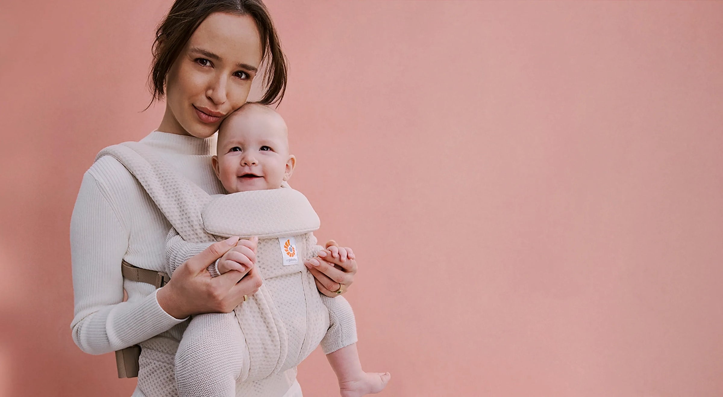 Eine Mutter, die ihr Baby in einer Ergobaby omni deluxe vor einem rosa Hintergrund hält